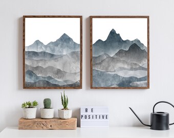 Puede incluir: Dos pinturas de acuarela enmarcadas de montañas azules y grises. Las pinturas están colgadas en una pared blanca sobre un estante de madera con tres suculentas en maceta y un letrero luminoso blanco que dice "SE POSITIVO".
