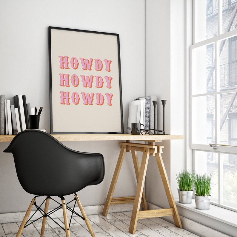 May include: A black framed print with the text "Howdy" repeated three times in pink and orange letters on a beige background. The print is hanging on a white wall above a wooden desk with a black chair and a wooden easel.