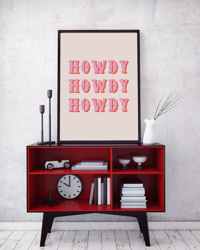 May include: A red shelf unit with black trim and three shelves. The top shelf has a framed print with the text "Howdy" repeated three times in pink letters. The middle shelf has a toy car, a clock, and books. The bottom shelf has more books and two small white cups.