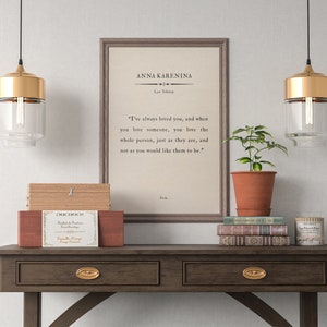 May include: A framed print of a quote from the novel "Anna Karenina" by Leo Tolstoy. The quote reads: "I've always loved you, and when you love someone, you love the whole person, just as they are, and not as you would like them to be." The print is hanging on a wall above a wooden table with two drawers. There are two gold pendant lights hanging above the table, and a potted plant is sitting on the table.