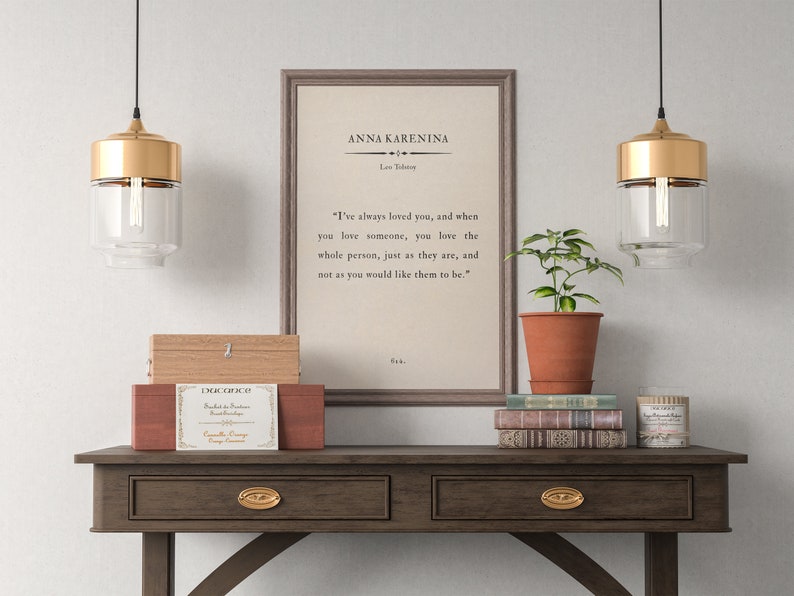 May include: A framed print of a quote from the novel "Anna Karenina" by Leo Tolstoy. The quote reads: "I've always loved you, and when you love someone, you love the whole person, just as they are, and not as you would like them to be." The print is hanging on a wall above a wooden table with two drawers. There are two gold pendant lights hanging above the table, and a potted plant is sitting on the table.