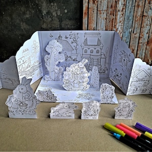 Op de afbeelding: Een zwart-witte papieren diorama van een fantasierijk dorp met konijnen, huizen en een fontein. De diorama bestaat uit verschillende panelen die kunnen worden gevouwen en geassembleerd. De diorama is klaar om te kleuren.