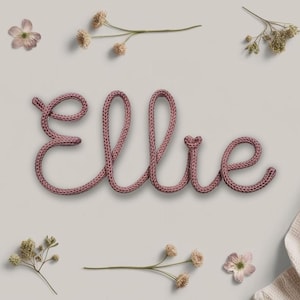 May include: A pink knitted word art piece spelling out the name "Ellie" surrounded by small white flowers and a white knitted blanket.