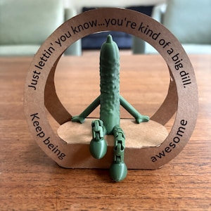 May include: A green pickle figurine with arms and legs, seated on a brown base. The figurine is surrounded by a circular cardboard frame with the text "Just lettin' you know...you're kind of a big dill. Keep being awesome."
