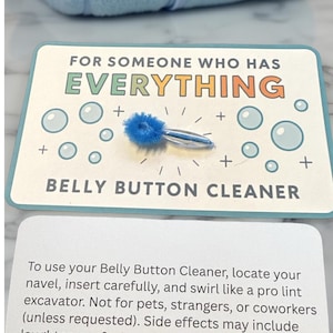 May include: A novelty gift set featuring a blue belly button cleaner with a fluffy head, presented on a card that reads "FOR SOMEONE WHO HAS EVERYTHING." The card has bubble illustrations. A blue towel is in the background.
