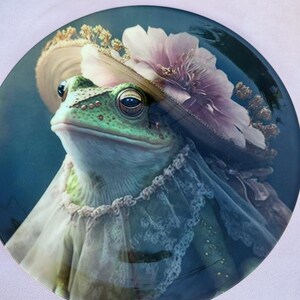 Può includere: Una rana verde che indossa un grande cappello color crema con un fiore rosa. La rana indossa un colletto di pizzo bianco e guarda in alto.