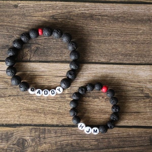 Puede incluir: Dos pulseras de cuentas de roca volcánica negra con cuentas de acento rojo. Una pulsera deletrea "DADDY" y la otra "HJM".