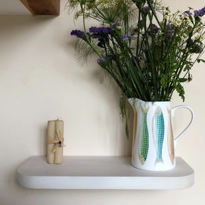 May include: A white ceramic jug with a colourful fish design sits on a white shelf. A bouquet of purple and green flowers is in the jug.