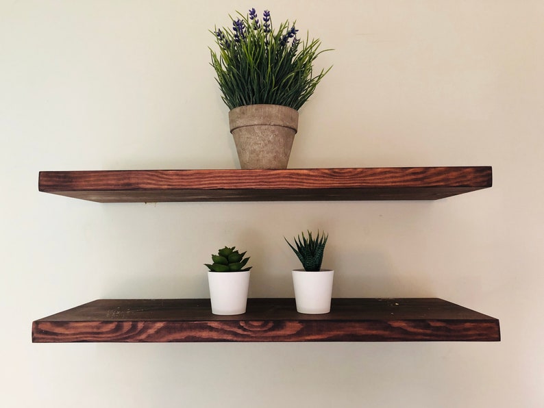May include: Two wooden shelves mounted on a wall, the top shelf has a potted plant with purple flowers and green leaves, the bottom shelf has two potted succulents, all in white pots.