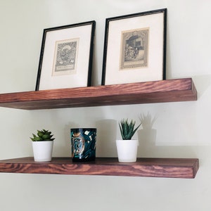 May include: Two wooden shelves mounted on a wall, with two framed black and white prints on the top shelf and two potted succulents and a decorative candle on the bottom shelf.