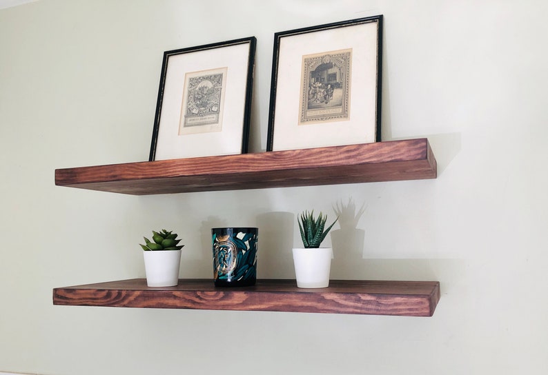 May include: Two wooden shelves mounted on a wall, with two framed black and white prints on the top shelf and two potted succulents and a decorative candle on the bottom shelf.
