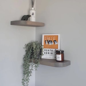 Op de afbeelding: Twee grijze hoekplanken met een plant in een pot, een boek getiteld "Ogata Korin Flock of Cranes", een kaars en een klein decoratief dierenbeeldje.