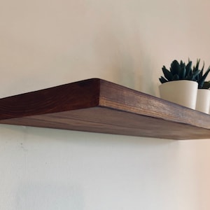 May include: A dark brown wooden shelf mounted on a white wall with two small potted plants on top.