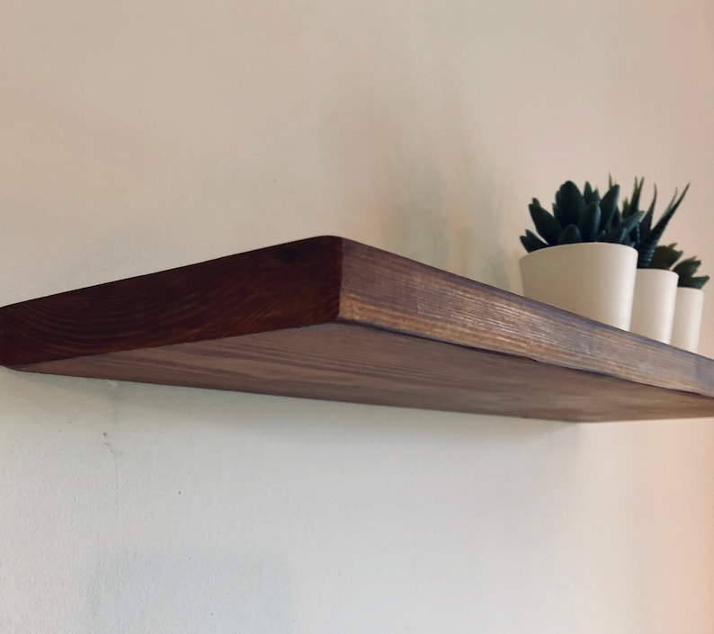 May include: A dark brown wooden shelf mounted on a white wall with two small potted plants on top.