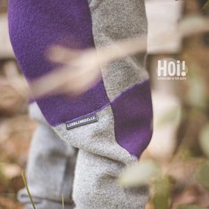 May include: Gray and purple pants featuring a color-blocked design with a textured fabric finish. A small rectangular label on the side displays the text LIEBLINGStil. The text Hoi! HANDMADE IN THE ALPS is visible on the upper right side.