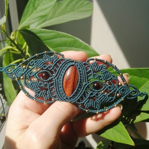May include: A teal macrame bracelet featuring a central oval wooden bead and smaller round beads. The intricate woven design incorporates two smaller, round, dark red beads. The bracelet is displayed against a backdrop of green leaves.