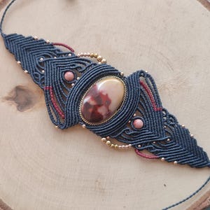 May include: A handcrafted macrame bracelet featuring a central oval gemstone in a brass setting. The bracelet is primarily navy blue with accents of red and small pink beads. Gold-colored beads and wooden beads complete the design.