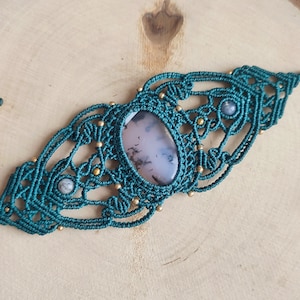 May include: Teal macrame bracelet featuring a large, oval, light pink and gray stone centerpiece. The macrame design includes small gold beads and two smaller gray stones. The bracelet is displayed on a wooden surface.
