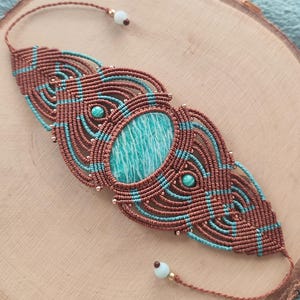 May include: A macrame bracelet with a central amazonite stone. The bracelet is made with brown and turquoise cords, and small gold beads. The woven design has a bohemian style. The bracelet is a unique piece of jewellery.