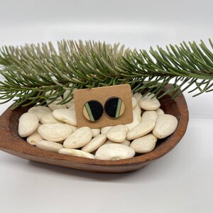 May include: A pair of round wooden earrings with black, green, and beige geometric patterns. The earrings are displayed on a small wooden bowl filled with white beans.