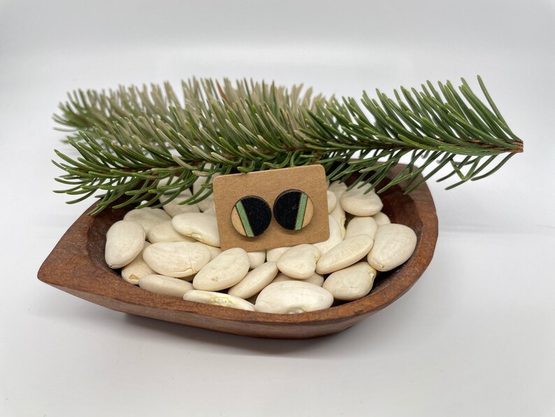 May include: A pair of round wooden earrings with black, green, and beige geometric patterns. The earrings are displayed on a small wooden bowl filled with white beans.
