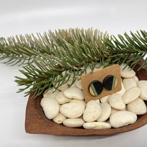May include: A pair of wooden earrings with black, green, and white geometric patterns. The earrings are displayed on a brown wooden bowl filled with white beans.