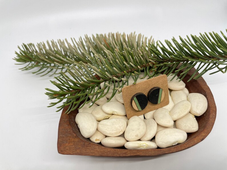 May include: A pair of wooden earrings with black, green, and white geometric patterns. The earrings are displayed on a brown wooden bowl filled with white beans.