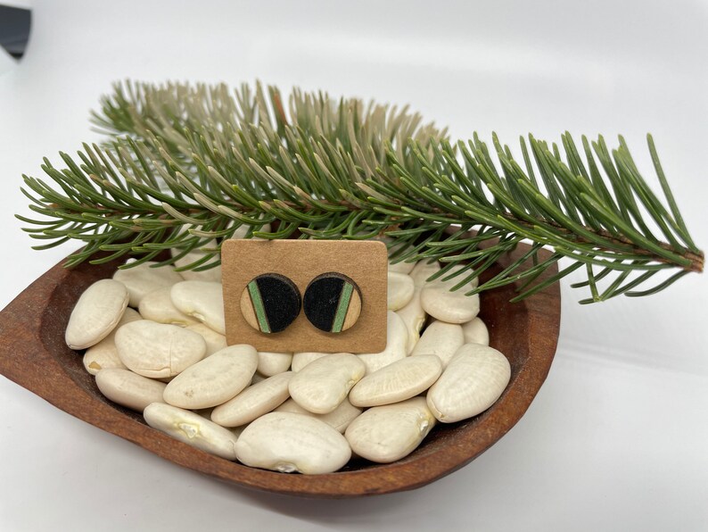 May include: A pair of small, round wooden earrings with black, green, and white stripes. The earrings are displayed on a brown wooden bowl filled with white beans and a green pine branch.