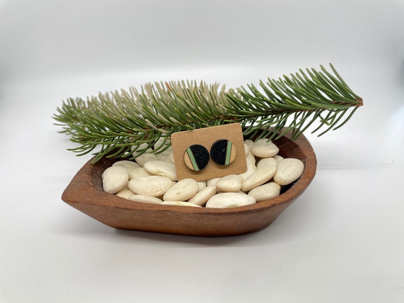 May include: A pair of small, round wooden earrings with black, green, and natural wood colors. The earrings are displayed on a small wooden bowl filled with white beans.