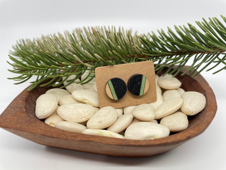 May include: A pair of wooden earrings with black, green, and natural wood stripes. The earrings are displayed on a small card with white beans in the background.