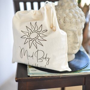 May include: A cream drawstring bag with a black sunburst design and the words "Mind Body made with soul." The bag is on a wooden chair next to a stone Buddha head and a vinyl record.