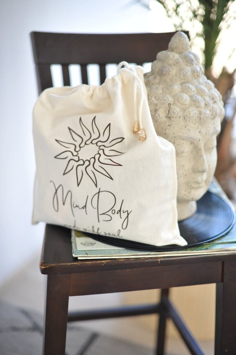 May include: A cream drawstring bag with a black sunburst design and the words "Mind Body made with soul." The bag is on a wooden chair next to a stone Buddha head and a vinyl record.