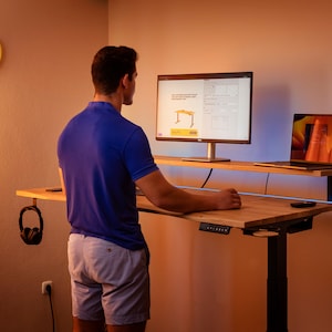 Peut inclure: Une personne debout devant un bureau debout avec un écran d'ordinateur et un ordinateur portable sur le bureau. Le bureau a un plateau en bois et un cadre en métal noir. L'écran d'ordinateur affiche un site Web pour un bureau debout.