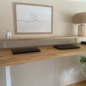 Könnte beinhalten: Ein Stehpult aus hellem Holz mit zwei Laptops auf der Oberfläche. Der Schreibtisch hat einen weißen Rahmen und ein weißes Regal über der Schreibtischoberfläche. Eine Lampe mit einem gewebten Schirm steht auf dem Regal.