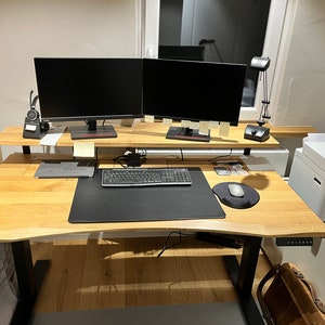 May include: A light wood standing desk with a black keyboard, mouse, and mousepad. Two black computer monitors are on a shelf above the desk. A white printer is on the right side of the desk.
