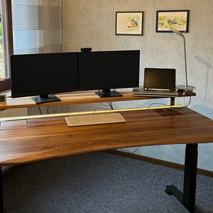 Peut inclure: Un bureau debout moderne avec un plateau en bois de noyer et des pieds en métal noir. Le bureau dispose d'une étagère intégrée pour un clavier et un support d'écran avec deux moniteurs. Il y a un ordinateur portable sur l'étagère, une lampe et une petite plante sur le bureau.