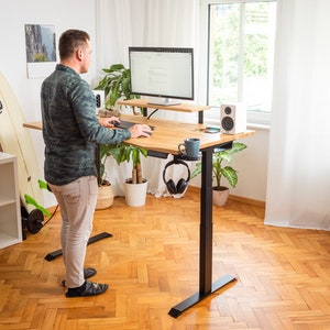 Op de afbeelding: Een in hoogte verstelbaar bureau met een licht houten blad en zwarte poten. Het bureau heeft een monitor, toetsenbord en een plank. Een persoon staat bij het bureau. Een koptelefoon hangt eraan. Het bureau staat in een kamer met een raam en een surfboard.