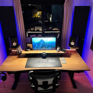 Può includere: Un ambiente di lavoro per ufficio a casa con una scrivania in legno regolabile in altezza, un monitor per computer, una tastiera, un mouse e due altoparlanti neri. La scrivania è illuminata da luci LED blu e le pareti sono decorate con pannelli acustici.