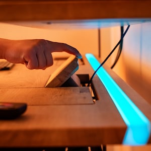 Op de afbeelding: Een houten bureau met een verborgen compartiment dat door een hand wordt geopend. Het bureau heeft een blauwe LED-lichtstrip die langs de rand loopt.