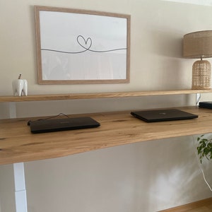 Op de afbeelding: Een staand bureau van licht hout met twee laptops op het oppervlak. Het bureau heeft een wit frame en een wit rek boven het bureauoppervlak. Een lamp met een geweven kap staat op het rek.