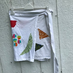 Peut inclure: Une jupe portefeuille blanche ornée de divers empiècements en tissu, dont un demi-cercle rouge, une forme de feuille verte, un motif floral et deux triangles orange et bleu marine. La jupe est en coton et possède un long lien de serrage blanc.