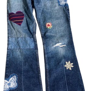 Puede incluir: Pantalones vaqueros azules con acabado desgastado, un parche de corazón morado, dos parches de mariposa blancos, un pequeño bordado floral y un dobladillo deshilachado con una banda decorativa ancha en tonos rojos, naranjas y morados con patrones geométricos.