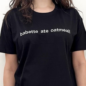 Puede incluir: Una camiseta negra con el texto "babette ate oatmeal" impreso en blanco en la parte delantera. La persona que lleva la camiseta también lleva leggings rosas.