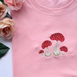 Könnte beinhalten: Hellrosa T-Shirt mit einer Stickerei von fünf roten Pilzen mit weißen Punkten. Das T-Shirt ist ordentlich gefaltet und liegt auf einer weißen Oberfläche. Rosa Rosen sind links oben zu sehen.