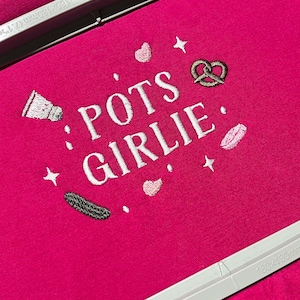 Puede incluir: Una camiseta rosa intenso con la frase bordada "POTS GIRLIE" en blanco, junto con imágenes bordadas de un salero, un pretzel, un pepinillo y corazones. La camiseta está sujeta en un bastidor de bordado plateado.