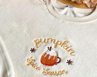 May include: Cream-colored sweatshirt with an embroidered design. The design features a pumpkin spice latte with the words "Pumpkin Spice Season" in gold thread. The sweatshirt has long sleeves and a crew neck. A small orange pumpkin and fall foliage are in the background.