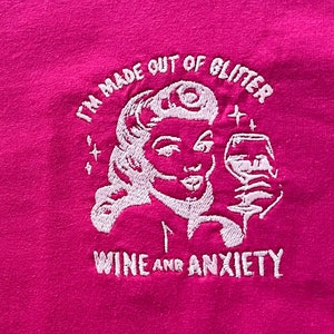 May include: Pink t-shirt with white embroidered text that reads "I'm made out of glitter, wine and anxiety". The design includes a stylized image of a woman with a glass of wine.