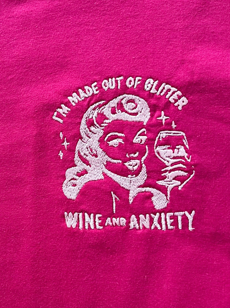 May include: Pink t-shirt with white embroidered text that reads "I'm made out of glitter, wine and anxiety". The design includes a stylized image of a woman with a glass of wine.