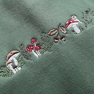May include: Green fabric with embroidered mushrooms and leaves in shades of brown, red, and white.