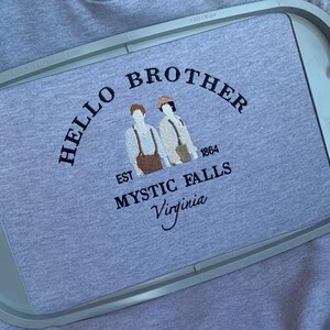 May include: A light gray t-shirt with the embroidered text "HELLO BROTHER" above two figures and the words "EST 1864 MYSTIC FALLS Virginia". The design is in dark brown thread.